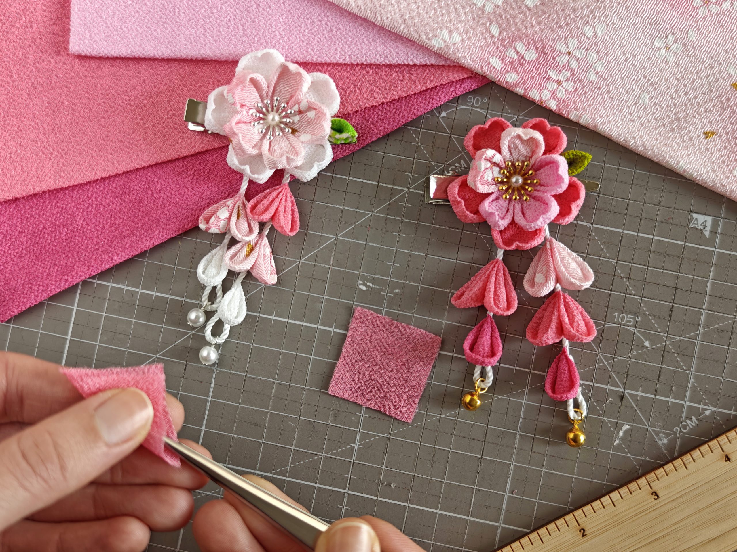 Due fermagli tsumami zaiku completati a forma di fiori di ciliegio (sakura), posizionati su un tappetino da taglio con griglia di misurazione. Il fermaglio a sinistra è rosa chiaro con petali pendenti e perle bianche, mentre quello a destra è rosa intenso con campanelle dorate e petali decorati. Sullo sfondo, pezzi di stoffa rosa e un righello in legno, con una mano che regge un pezzo di tessuto rosa e una pinza.